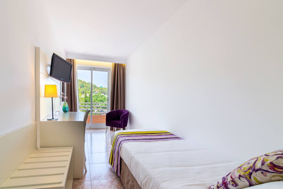 A hotel room with a bed, a desk with a chair, a TV, and a window with curtains.