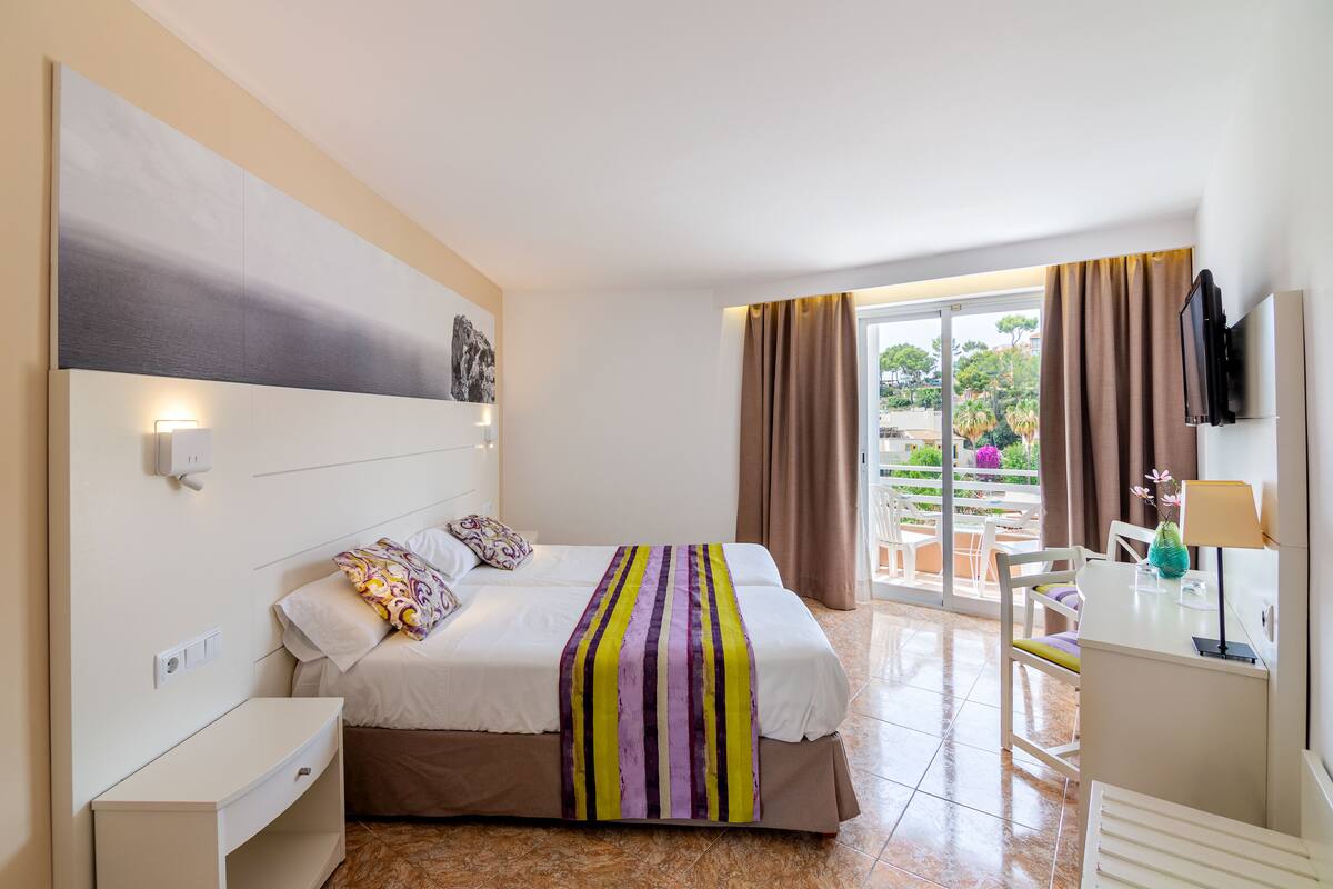 A hotel room with a bed, bedside table, and a desk with a lamp. There is a window with curtains and a view of the outside.