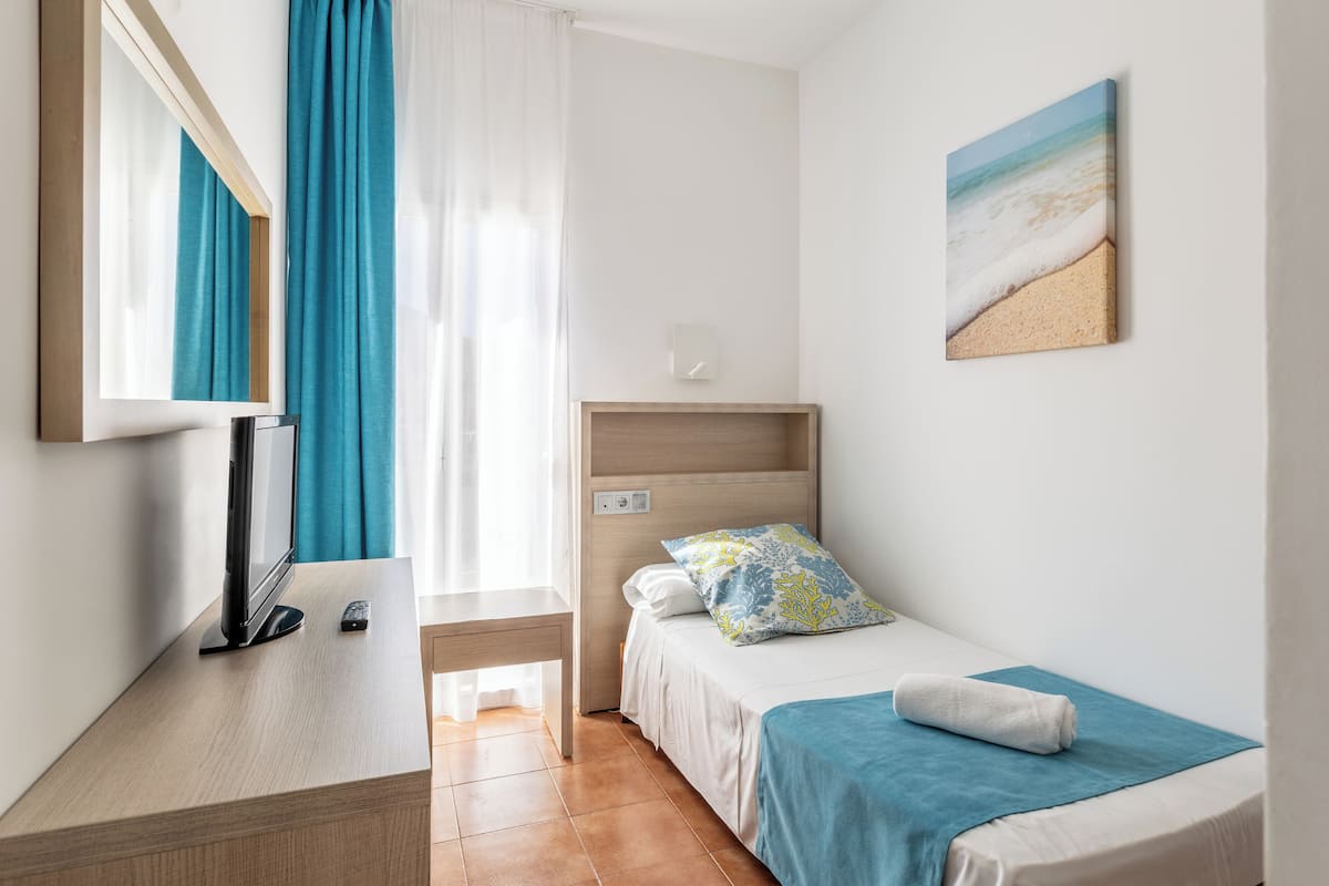 A hotel room with a bed, a desk with a TV, a window with curtains, and a painting on the wall.