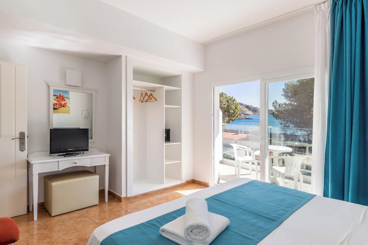 A hotel room with a bed, a desk with a TV, a wardrobe, and a balcony with a view of the sea.