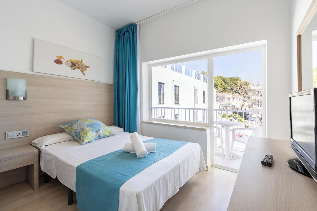 A hotel room with a bed, a desk with a TV, a window with a view of buildings, and a wall-mounted artwork.