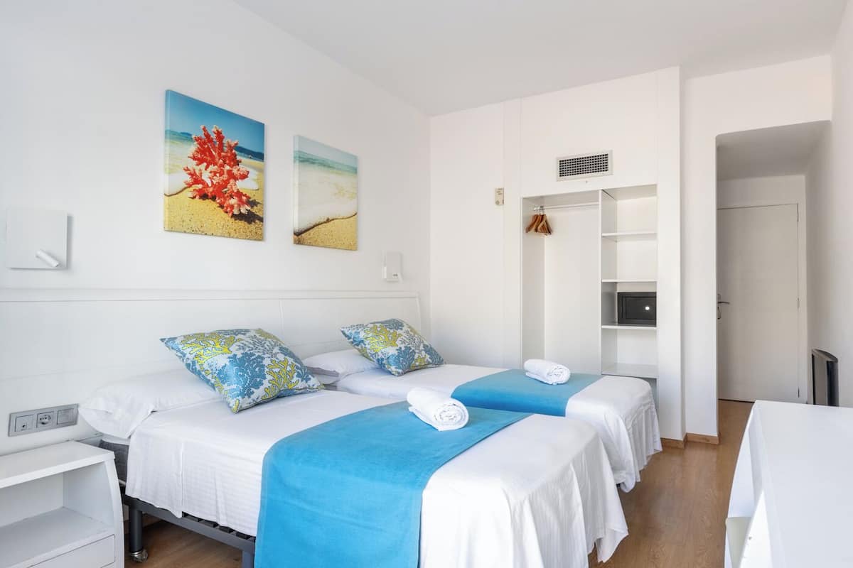 A hotel room with two beds, each with a blue and white bedspread, and matching pillows. There are two paintings on the wall, a white wall-mounted heater, and a door leading to another room.