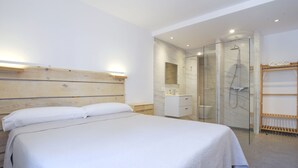A modern bedroom with a large bed, a wooden headboard, a glass-enclosed shower, and a small bathroom with a mirror and sink. - In the middle of Pollensa Bay (Apto Garrover A) (Port de Pollença)
