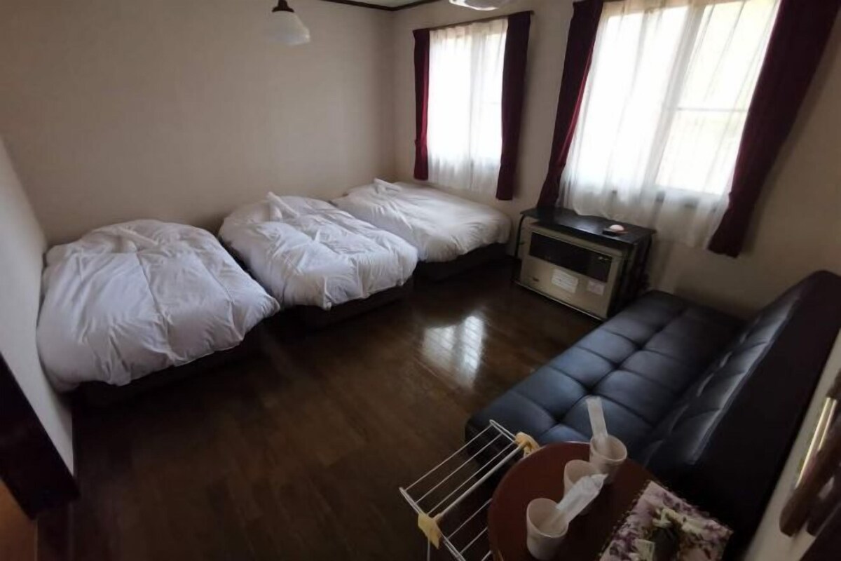 Una habitación con dos camas individuales, un sofá, una mesita con una taza y una cuchara, y una ventana con cortinas.