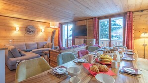 Et hyggeligt interiør i en hytte med et spisebord dækket til morgenmad, trævægge og et stort vindue med udsigt over et sneklædt landskab.