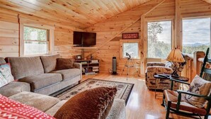 Ein gemütliches Blockhaus-Interieur mit Holzbalkendecke, Flachbildfernseher, einer Couch, einem gemusterten Sessel und einem Teppich.