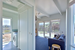 Ein Waschbereich mit Waschmaschine, ein blau teppichausgelegter Raum inklusive Sofa und Hocker, sowie große Fenster mit Blick ins Freie.