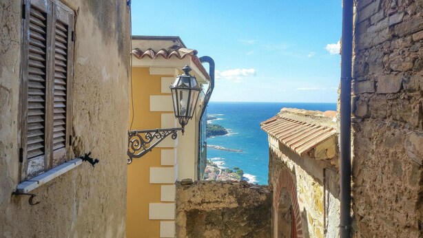 A narrow street with a view of the sea, a lamp post, and a building with a tiled roof.