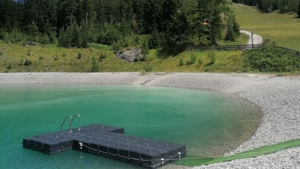 Un lago turquesa cristalino con un muelle, rodeado por una playa de guijarros y un bosque denso, con montañas al fondo.