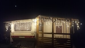 Una caravana pequeña y blanca con un porche de madera, decorada con luces colgantes.