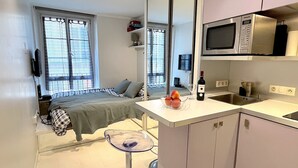 Habitació compacte amb un llit, una taula de nit i una finestra petita. A prop, hi ha un petit espai cuina amb microones, rentapeus i un plat de fruita.