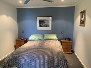 A bedroom with a bed, two bedside tables, a ceiling fan, a framed picture on the wall, and a small table with a lamp.