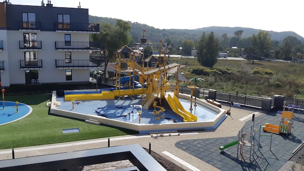 Un terrain de jeux équipé de toboggans et de structures d’escalade, entouré d’une piscine et d’un bâtiment doté de balcons.