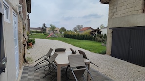 Une terrasse avec une table et des chaises, un chemin de gravier et une vue sur une aire de jeux et des maisons au loin.