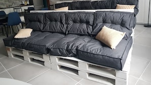 Un canapé noir avec des coussins, posé sur une base blanche, agrémenté de plusieurs oreillers, se trouve dans une pièce dont l’arrière-plan dévoile une cuisine et un escalier en bois.