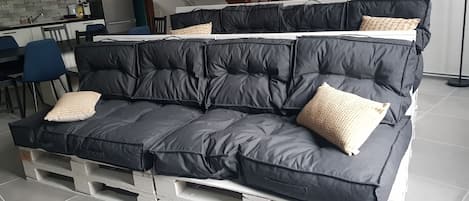 Un sofá negro con cojines sobre una base blanca, situado en una habitación con una cocina al fondo y una escalera de madera.