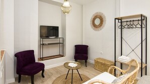 Un salon moderne avec un téléviseur, deux fauteuils violets, un canapé en osier, un porte-manteau suspendu et un miroir décoratif.