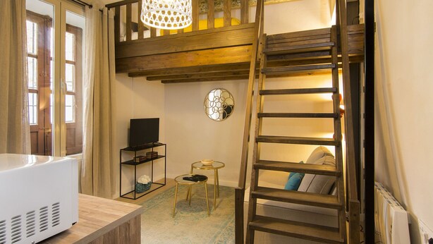 Una habitación compacta con cama estilo altillo, un televisor sobre un soporte, una pequeña mesa redonda y una escalera para acceder al altillo.