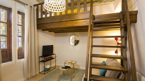 Un habitación tipo loft con una escalera de madera, un televisor sobre un soporte negro, una mesita redonda con una lámpara y una alfombra estampada.