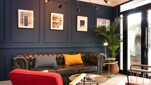 Pared azul oscuro con marcos, un sofá de cuero con cojines amarillo y gris, un sillón rojo, una mesita redonda con una revista y una planta en maceta.