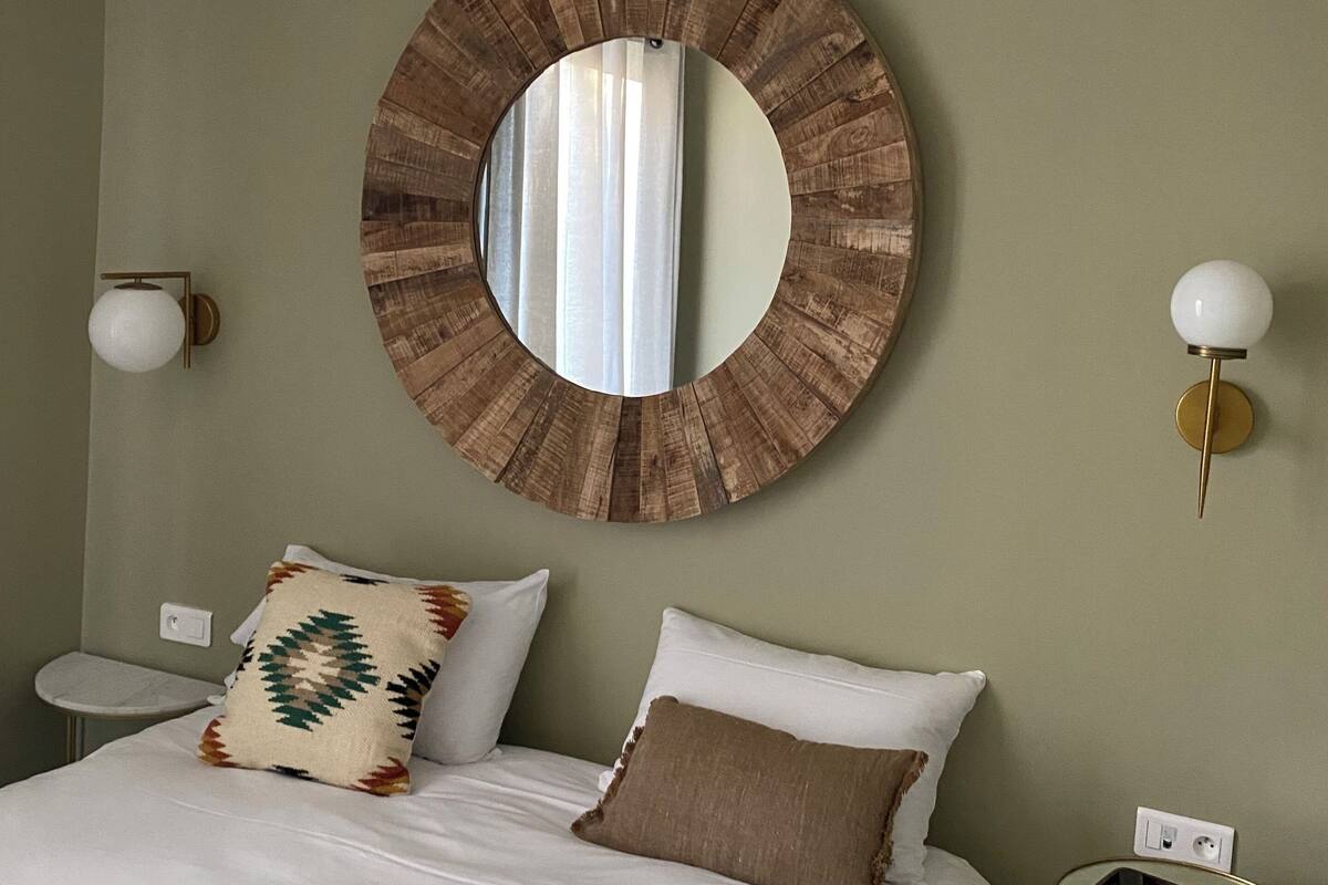 A bedroom with a round mirror, a bed with white linens, two pillows, a bedside table, and a small round table with a lamp.