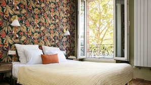 Un dormitorio con cama, almohadas blancas y anaranjadas, manta color crema, papel tapiz floral, ventana con vista a un árbol y balcón con barandilla de metal.