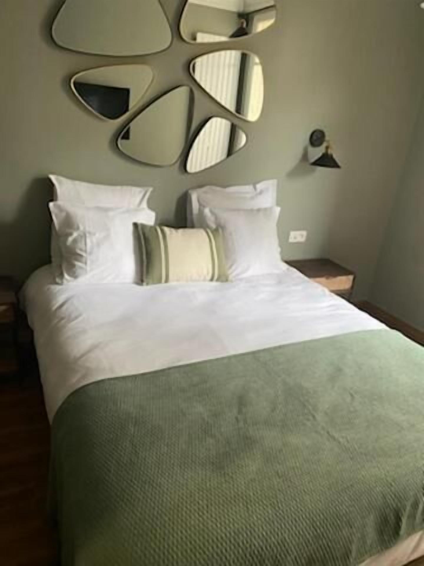 A neatly made bed with white linens and a green throw blanket, a bedside lamp, and a wall-mounted mirror.
