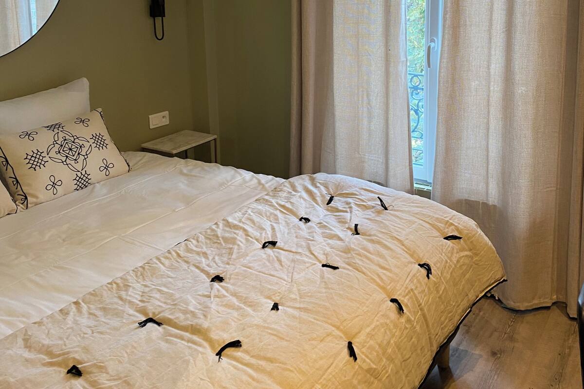 A bedroom with a large bed, a bedside table, a wall-mounted phone, and a window with curtains.