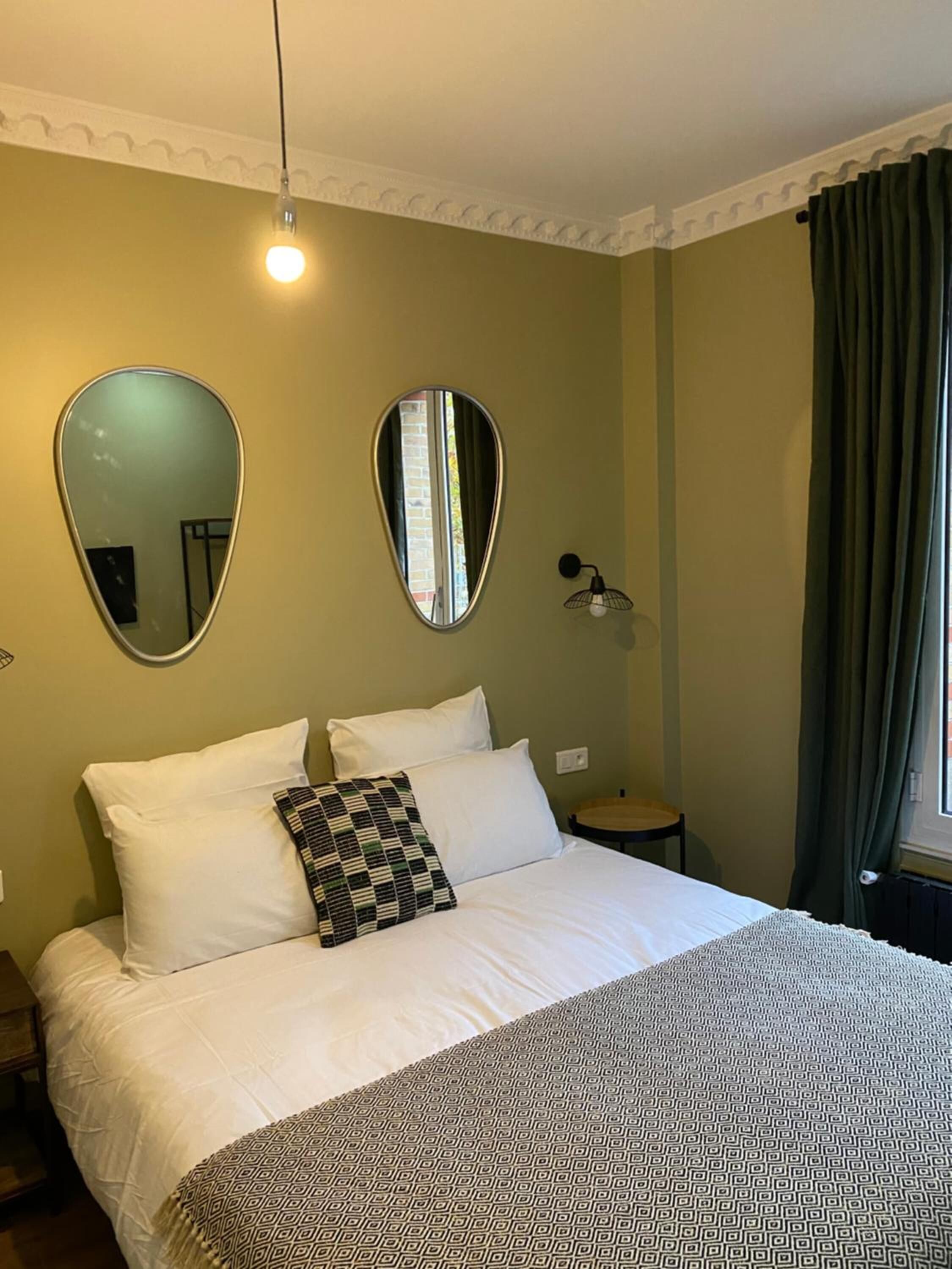A neatly made bed with white linens and patterned pillows, a large mirror on the wall, and a small table with a lamp beside it.