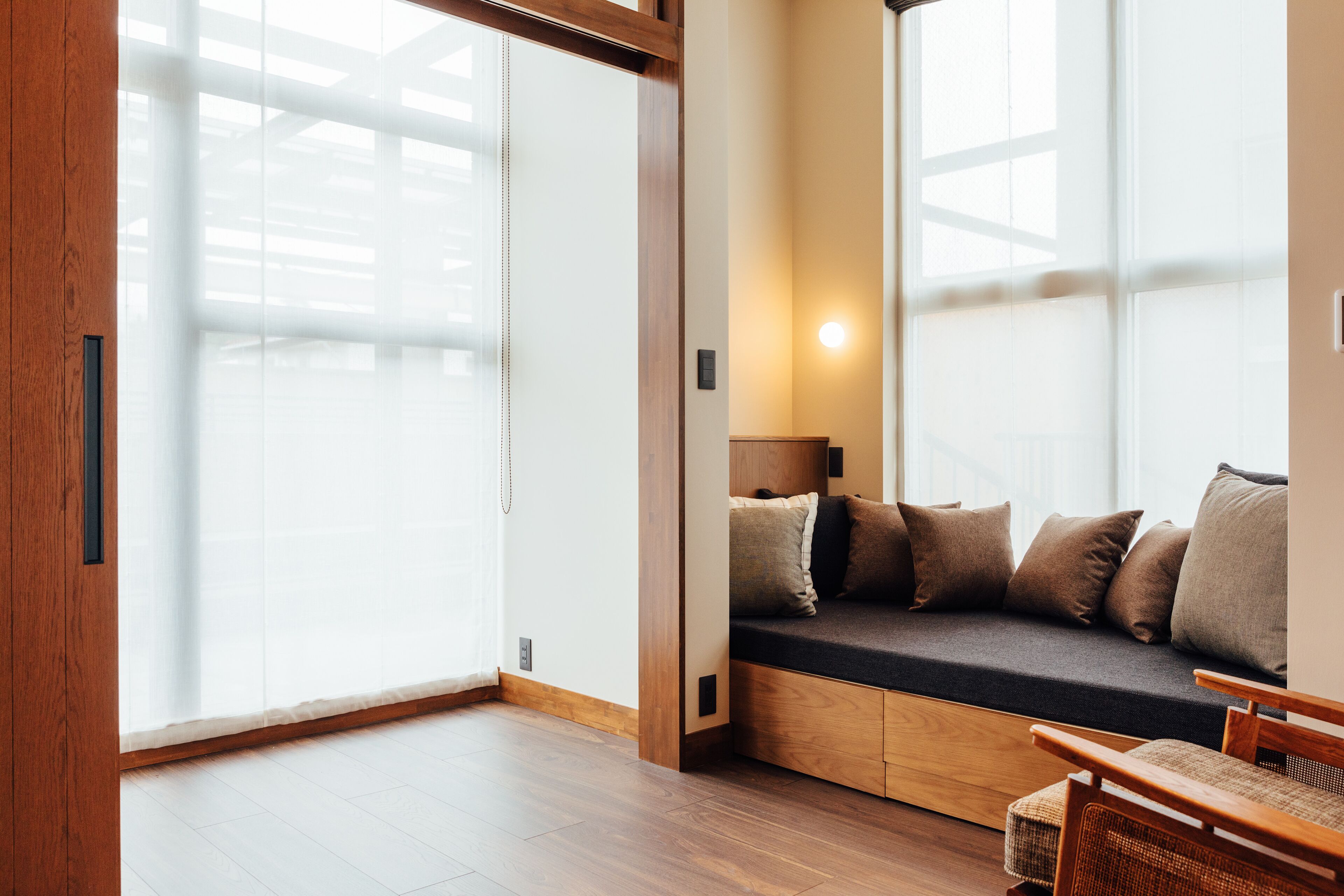 A modern room with a wooden floor, a built-in bench with cushions, a wall-mounted lamp, and large windows with frosted glass.