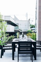 Un área de comedor al aire libre con una mesa preparada para cuatro, rodeada de sillas negras, y un edificio moderno al fondo.