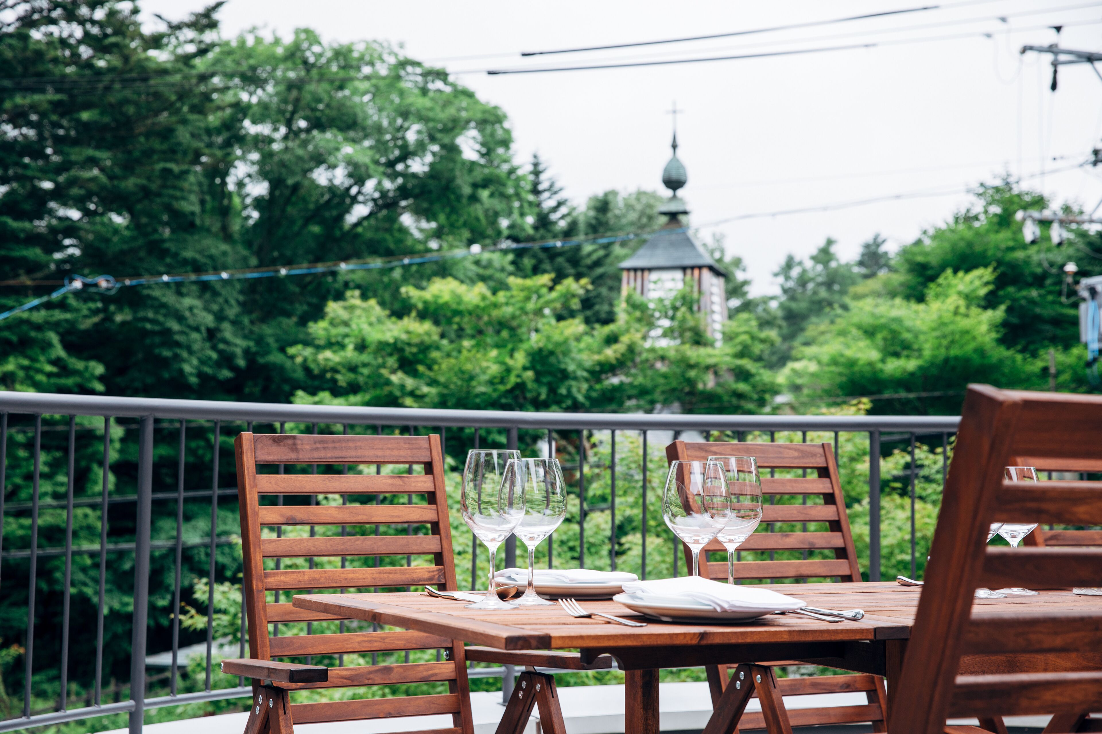 A wooden table set with plates, cutlery, and wine glasses on a balcony with a view of trees and a distant building.
