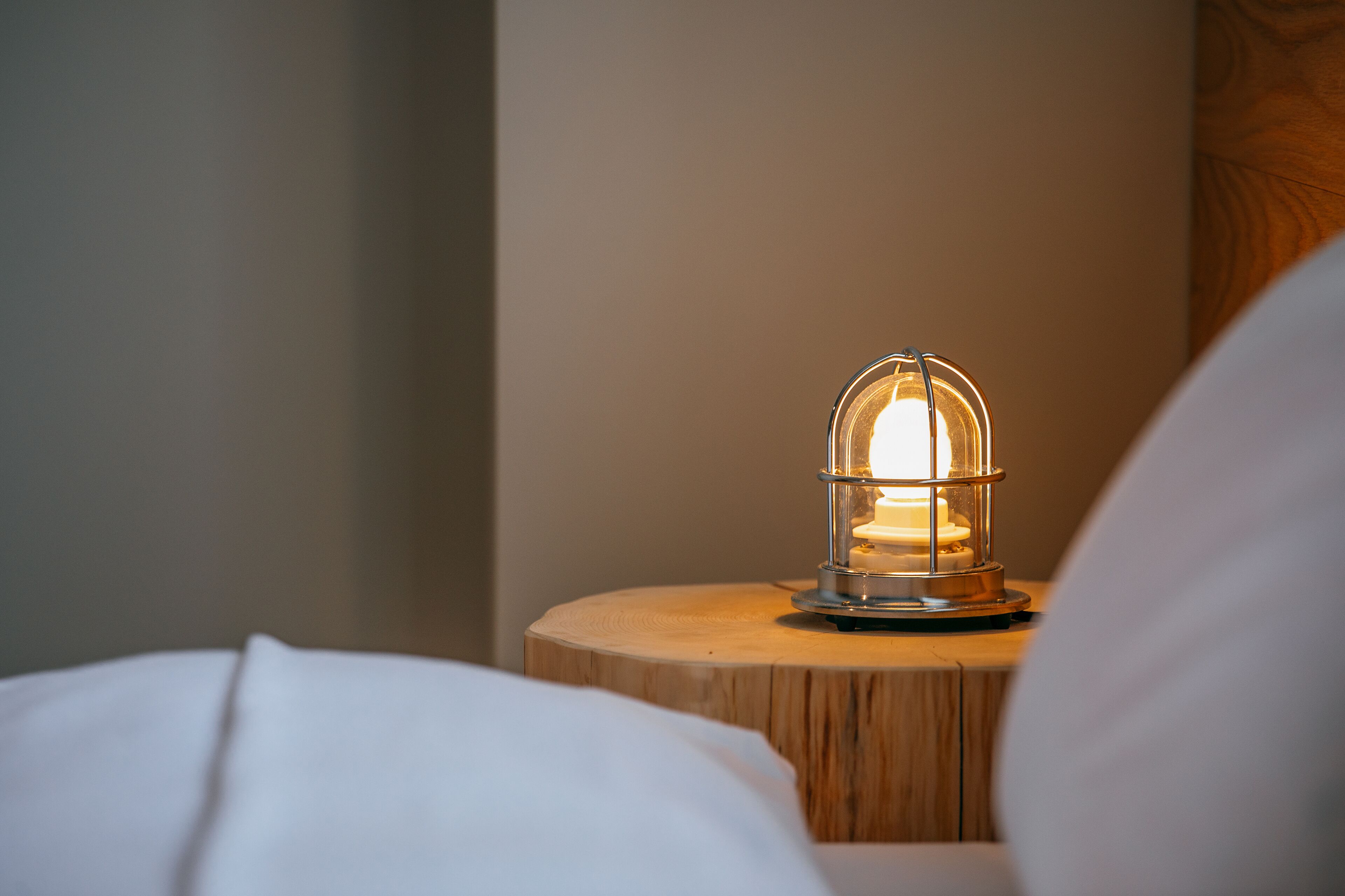 A bed with white bedding and a round wooden nightstand with a lit lamp.