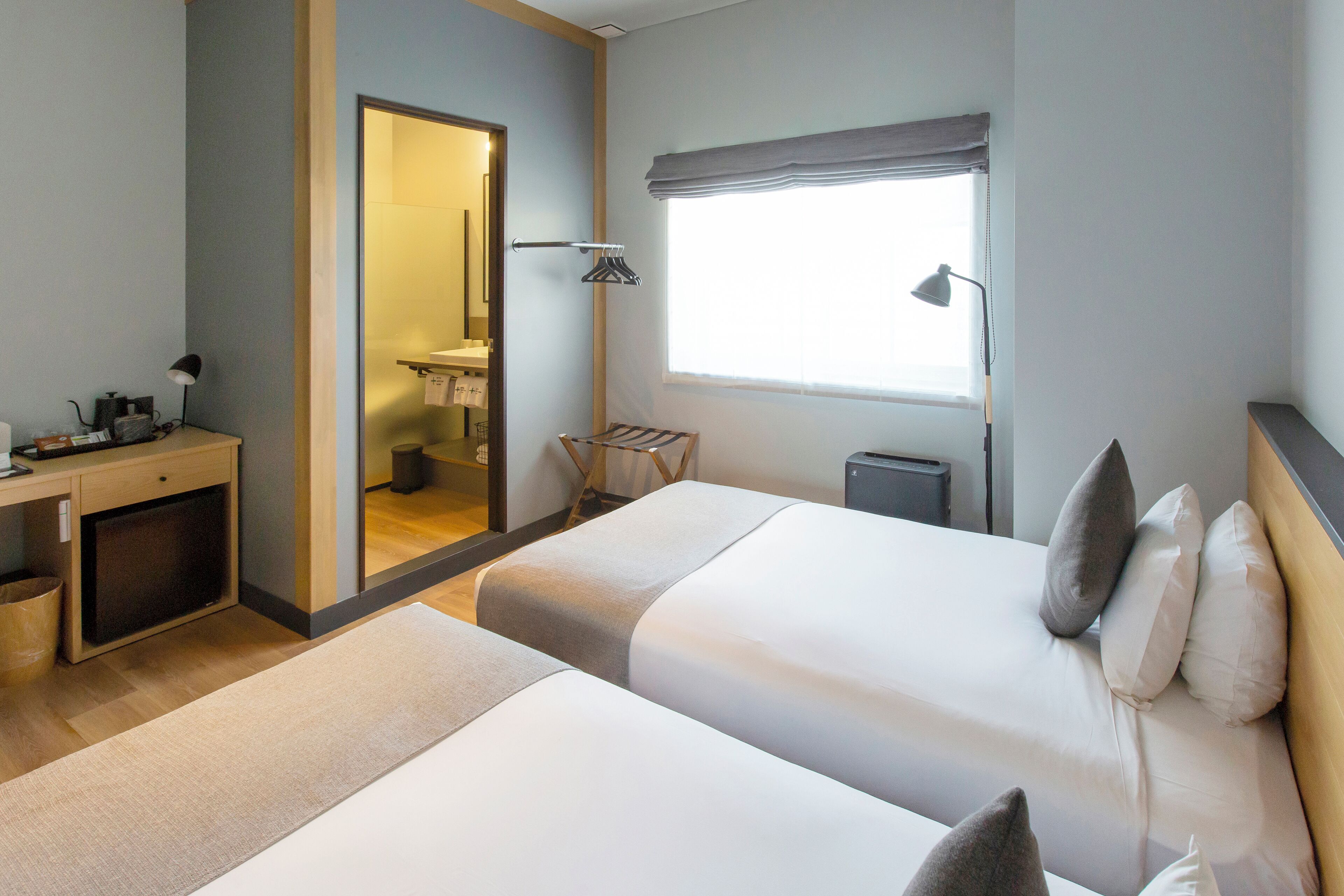 A hotel room with two beds, a desk, a chair, a window with a view, and a bathroom visible through an open door.