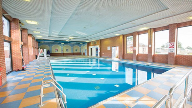 En inomhuspool med ett rutmönstrat klinkergolv, kristallklart blått vatten och utsikt över omgivningen genom stora fönster.