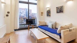 A living room with a sofa, a bench, a TV, and a large window with curtains. - Beautiful 1 Bedroom Apartment in Shepherd's Bush (London)