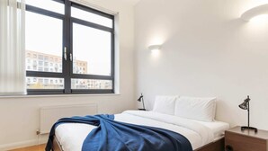 A bedroom with a bed, a nightstand, a lamp, a window, and a view of buildings outside. - Beautiful 1 Bedroom Apartment in Shepherd's Bush (London)