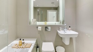 Une salle de bain moderne avec un lavabo blanc, un miroir et des toilettes. On peut voir des articles de toilette sur le comptoir et une pomme de douche qui se reflète dans le miroir.