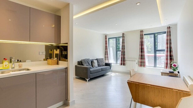 Une cuisine moderne dotée d’un îlot central intégré, d’un canapé gris et d’une table à manger en bois, le tout dans un espace de vie lumineux avec de grandes fenêtres.