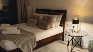 A neatly made bed with a wooden headboard, a bedside table with a lamp, and a large window with curtains. - Modern 1 Bedroom Apartment Near Canary Wharf With Balcony (London)