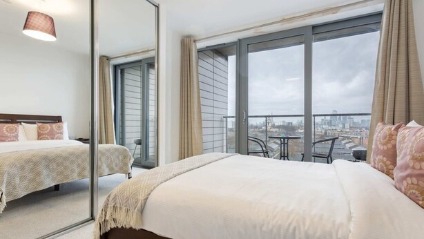 A hotel room with a large bed, a second smaller bed, a balcony with a city view, and a mirror reflecting the room. - Modern 1 Bedroom Apartment Near Canary Wharf With Balcony (London)