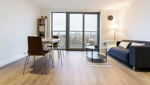 A modern living room with a dining area, large windows with a city view, a sofa, and a lamp. - Modern 1 Bedroom Apartment Near Canary Wharf With Balcony (London)