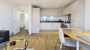 A modern kitchen with white cabinets, a wooden dining table, and a glass coffee table. - Modern 1 Bedroom Apartment Near Canary Wharf With Balcony (London)