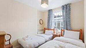 Et hotelværelse med to senge, et trænatbord, en stol, et vindue med mønstrede gardiner og udsigt til bygninger.
