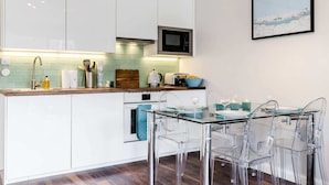 A modern kitchen with white cabinets, a microwave, a glass dining table with clear chairs, and a framed picture on the wall. - Lovely 2 Bedroom Apartment With Garden, South Bank Westminster (London)
