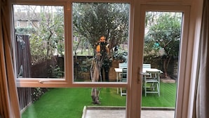 A room with a large sliding glass door leading to a backyard with a lawn, a tree, and a brick wall. - Lovely 2 Bedroom Apartment With Garden, South Bank Westminster (London)