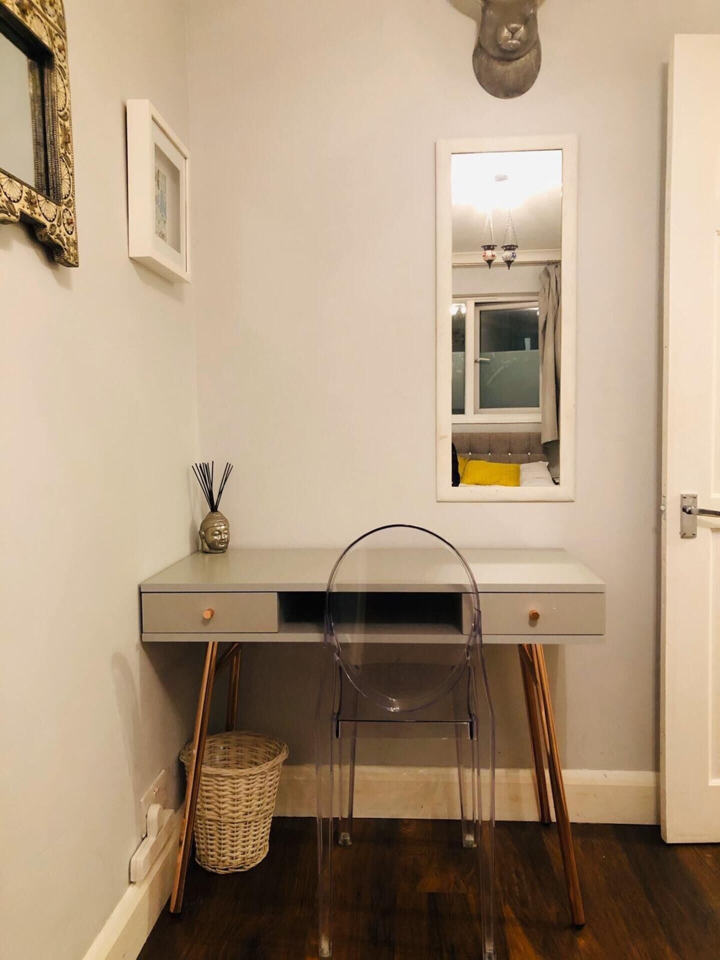 A small, modern desk with a clear chair in front, a basket, and a framed picture on the wall.