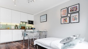 A modern bedroom with a bed, a desk, and chairs, a kitchen area with white cabinets, and four framed artworks on the wall. - Lovely 2 Bedroom Apartment With Garden, South Bank Westminster (London)