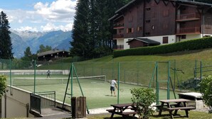 A tennis court with a net, surrounded by a green fence, and a building with a balcony in the background. - Apartment Combloux sleeps 4 at the foot of the slopes (Combloux)