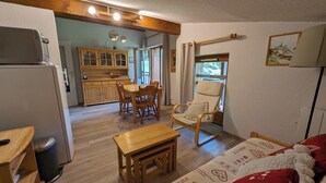 A cozy living room with a wooden table, a chair, and a sofa. There is a kitchen area with wooden cabinets and a refrigerator. - Apartment Combloux sleeps 4 at the foot of the slopes (Combloux)
